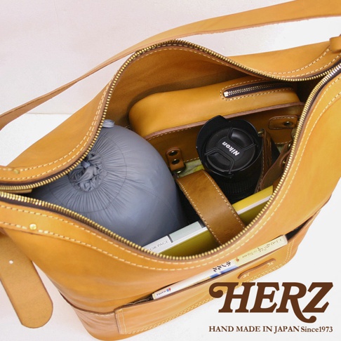 駝色皮革單肩波士頓包，內部裝有灰色睡袋、黃色內袋、黑色相機鏡頭蓋及書籍。包包以厚實皮革製成，配有拉鍊開合及皮革肩帶，走線清晰。