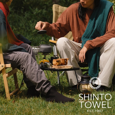 兩位人士在戶外草地上，一人手持咖啡濾杯，桌上擺放著茶壺、咖啡杯與暖色調的燈籠。其中一人身上披著一條深綠色毛毯，展現溫馨的露營氛圍。