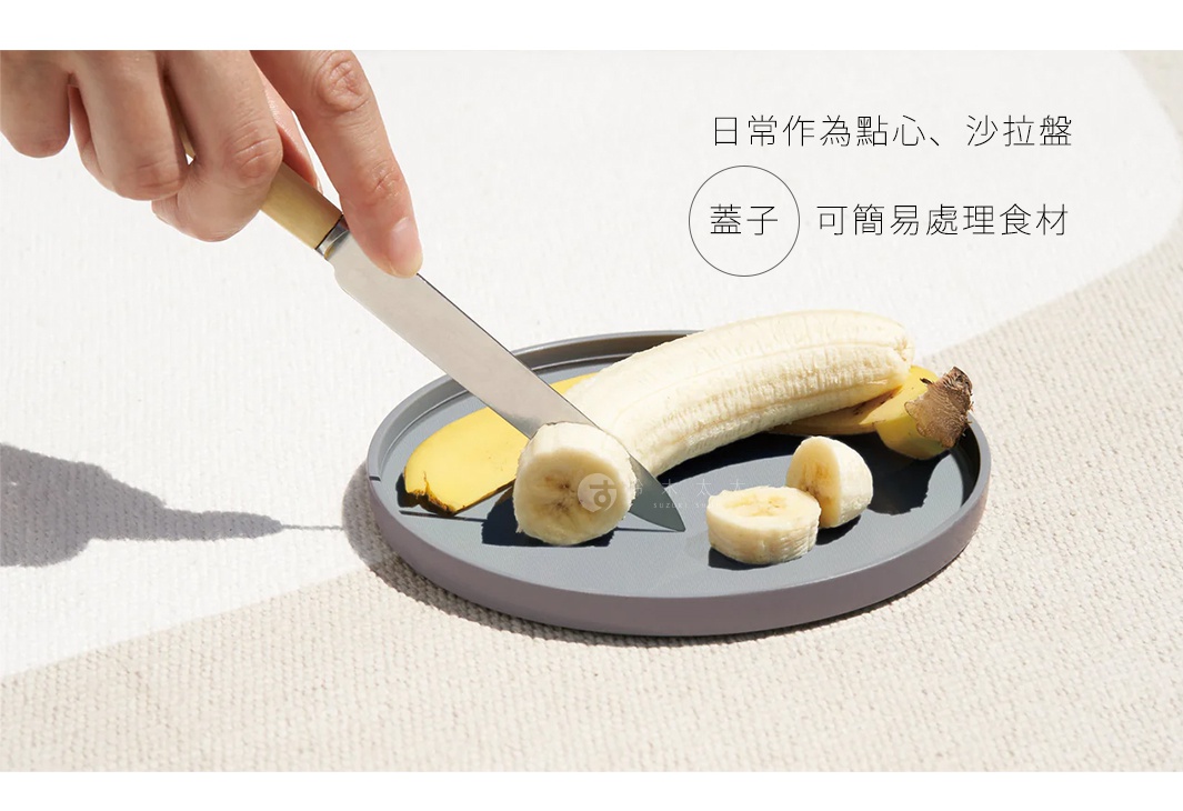 灰色圓形防摔餐盤上，一把木柄不鏽鋼刀正在切香蕉，盤中已有香蕉片和一根未切的香蕉。