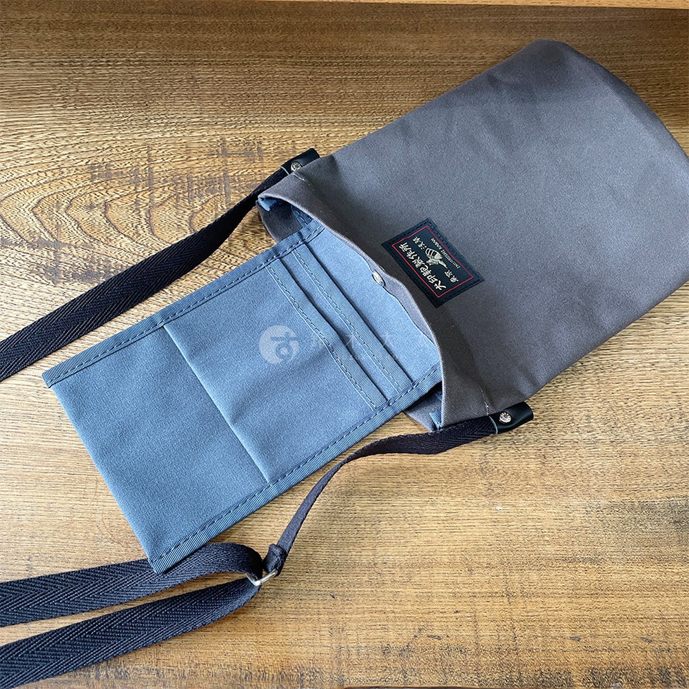 灰色帆布平板收納側背包，內有淺藍色卡夾層，附有黑色斜揹帶，放置於木紋桌面上。