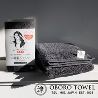 深灰色毛巾，觸感蓬鬆，質地柔軟，疊放在白色表面上，旁邊是產品包裝盒，上面印有手繪圖案和日文說明。