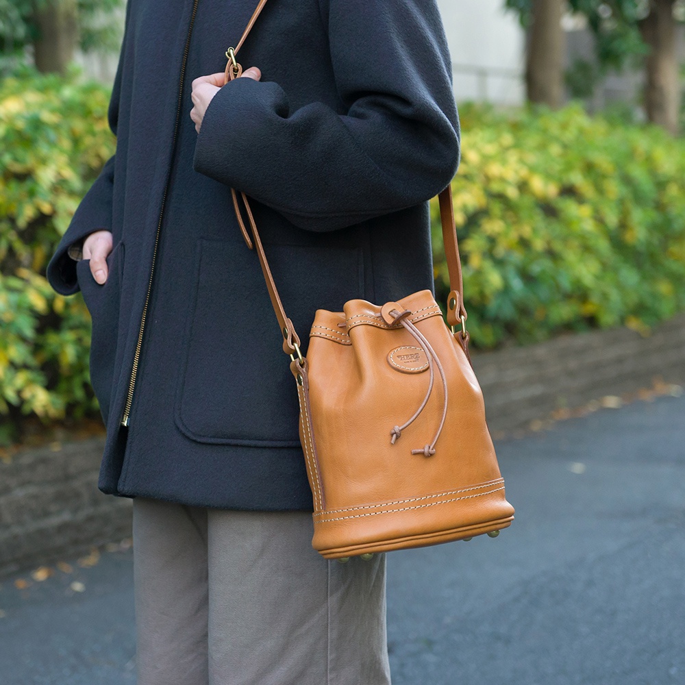 棕褐色皮革束口單肩包，有著經典的圓潤包身設計，束帶和縫線細節清晰可見，搭配素色外套和褲子。