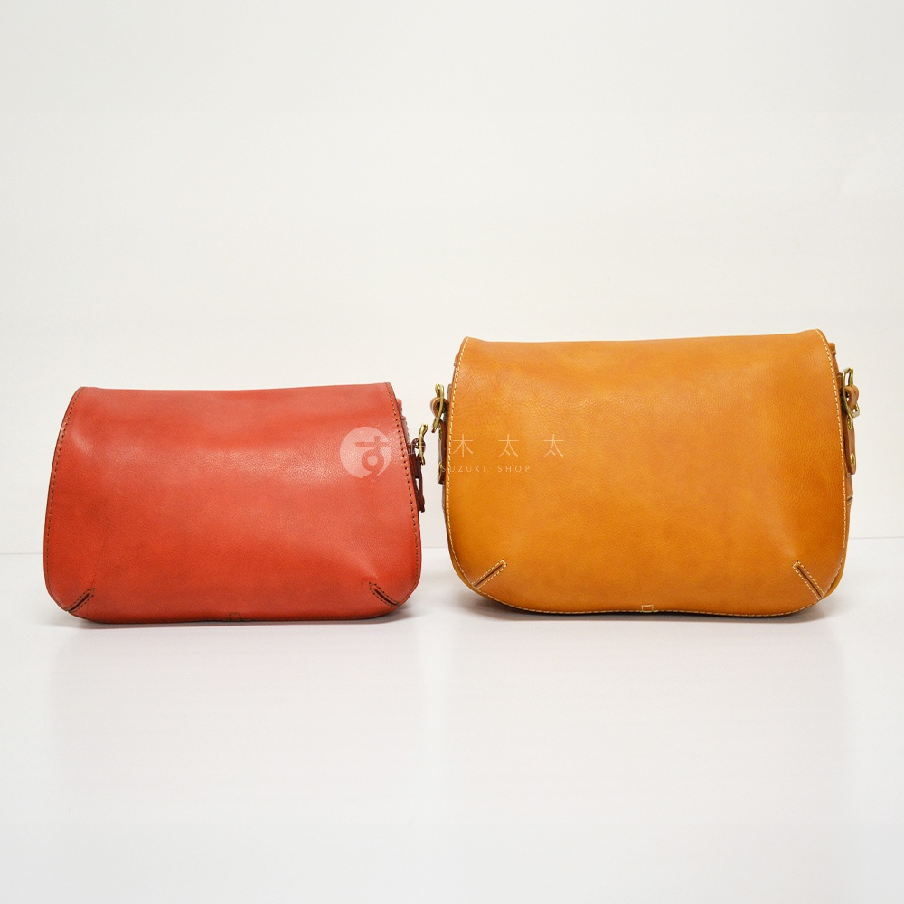 兩個職人手作皮革復古翻蓋郵差包，一個為紅色，另一個為橘黃色，皆為真皮材質，有著精緻的車縫線與金屬扣環。