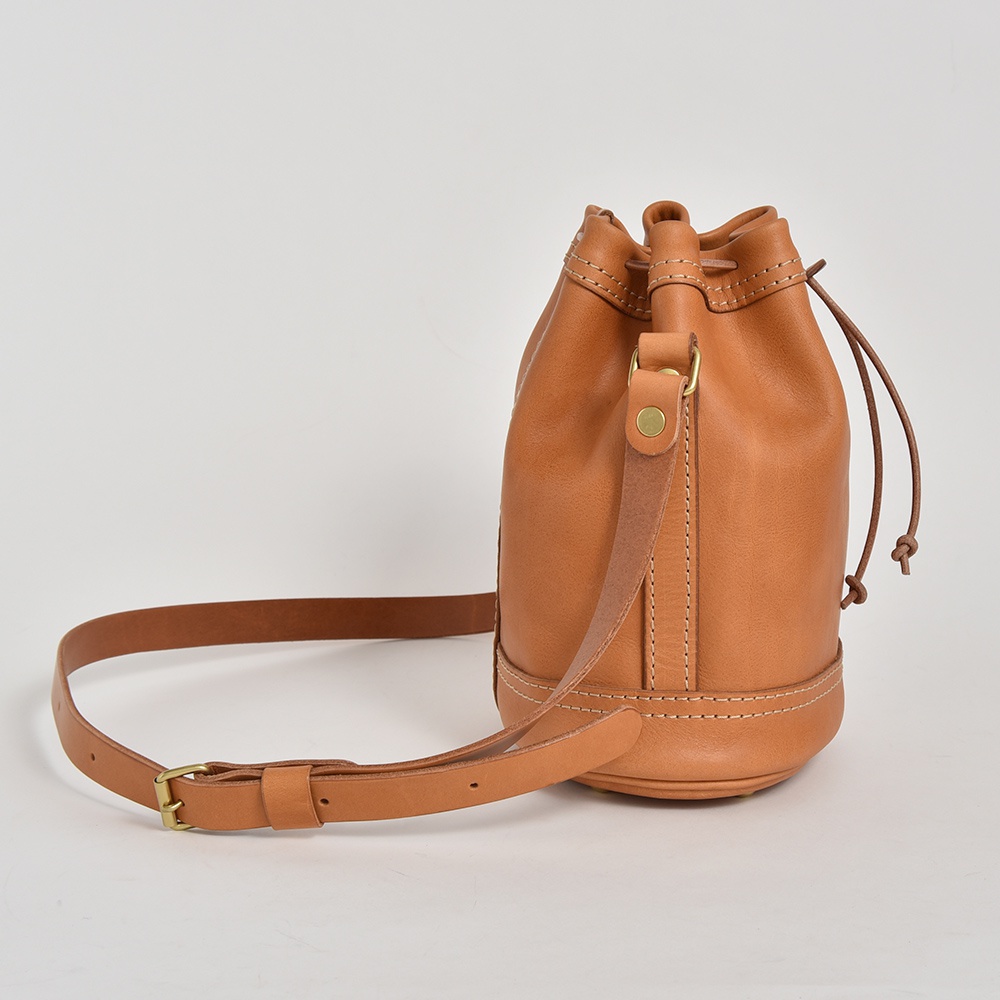 職人手作皮革束口單肩包，採用淺棕色皮革製成，設有束繩開口、可調節式單肩背帶及金屬釦環，包身結構穩固，車縫線精細。