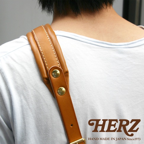 棕色皮革加厚減壓肩背帶，配有兩顆黃銅色鉚釘及金屬扣環，手工縫線精緻，展現職人質感。