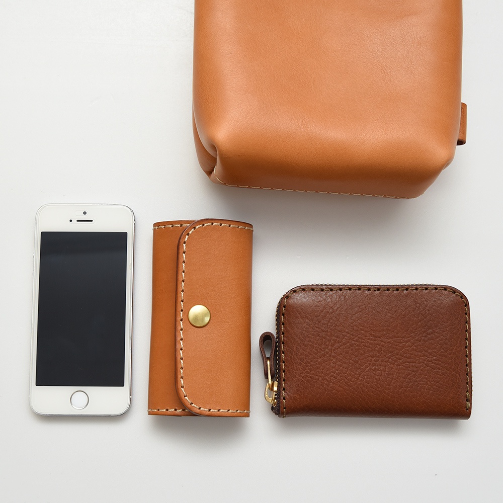 職人手作皮革多用途工作用腰袋 E-27，展示了淺棕色和深棕色的皮製配件，包含一個手機、一個帶有金色銅釦的鑰匙包，以及一個拉鍊零錢包。