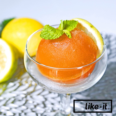 玻璃杯中盛著一顆橙色圓形冰球，頂部裝飾著綠色薄荷葉，旁邊擺放著檸檬片。背景是模糊的黃色檸檬和白色紋理。