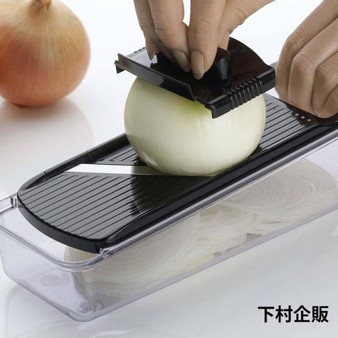 一雙手正使用下村企販品牌的黑色切片器刨切洋蔥，切好的薄片整齊地堆疊在下方的透明收納盒中，背景另有一顆完整的洋蔥。