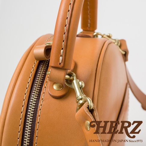 HERZ 品牌標誌與「HAND MADE IN JAPAN Since1973」字樣，搭配經典棕色皮革手提包的拉鍊與金屬掛鉤細節