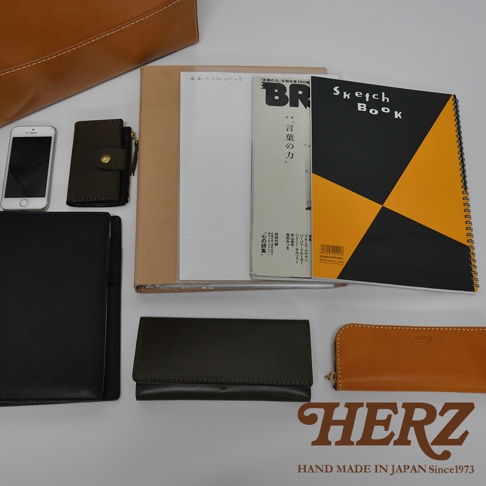 職人手作皮革直式托特包 T-44 L，展示了包含一個手機、一個深綠色皮革零錢包、一個棕色皮革長夾、一個黑色皮革夾、一本筆記本、一本雜誌和一本黑色黃色幾何圖案的 Sketch Book。