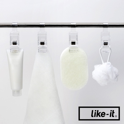 四個透明塑膠掛夾夾在金屬桿上，分別夾著一管白色乳液、一條白色毛巾、一個淺綠色浴球和一個白色網狀沐浴球。