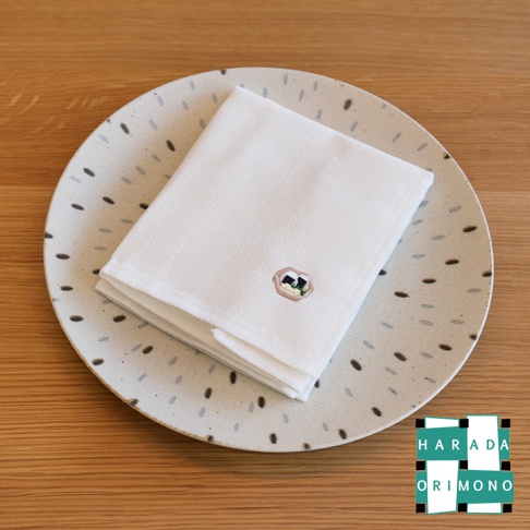 一張疊好的白色八重紗擦拭巾，上面繡有小巧的壽司圖案，放置於一個帶有斑點圖案的淺色盤子上，盤子置於木質桌面。