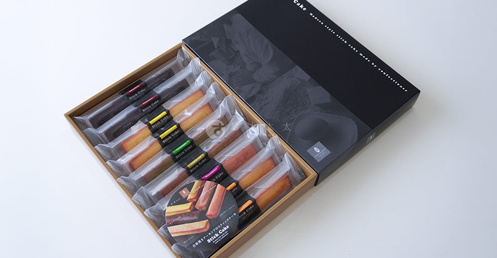 模式B： 一個木製禮盒裝著井桁堂的長條費南雪蛋糕，蛋糕外層為獨立透明包裝，上面印有Stick Cake字樣，禮盒蓋為黑色，上面印有品牌標誌和現代風格的文字。