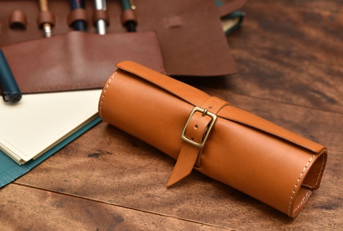 焦糖色皮革捲捲筆袋，配有黃銅扣環及皮帶，側邊及邊緣有白色車線。筆袋旁攤開的棕色皮革筆簾內有數支筆，左側有一疊白色紙張及一支藍色鋼筆。