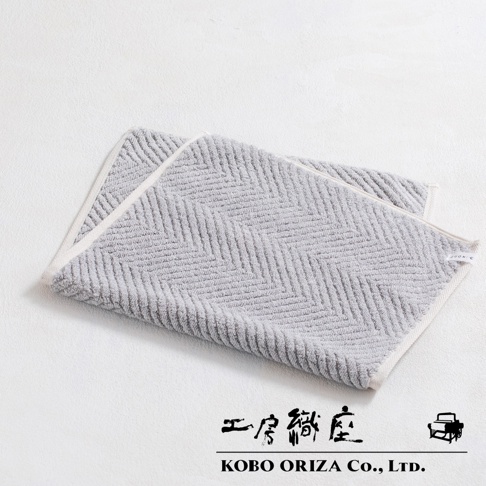 兩條淺灰色棉麻混紡人字紋快乾浴墊，一條疊在另一條上面，邊緣有白色滾邊。