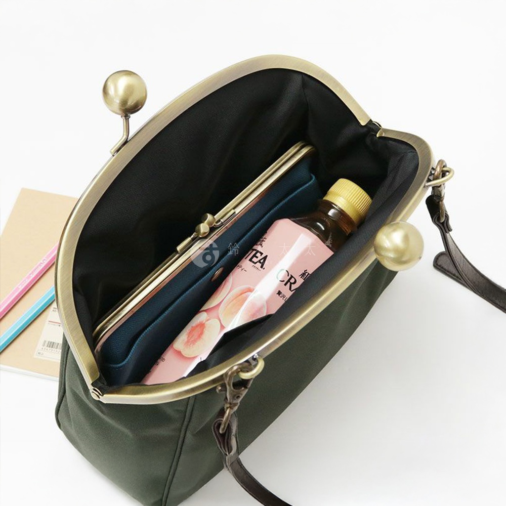 一個綠色帆布斜背包，打開後露出內部，裡面有一個錢包和一瓶飲料，包包配有古銅色金屬開扣和肩帶。