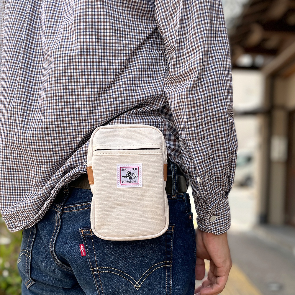 石洗帆布斜背手機包，淺米色，正面有方形標籤，帶有圖案和文字，包包側邊有皮質掛環，可掛於腰間。
