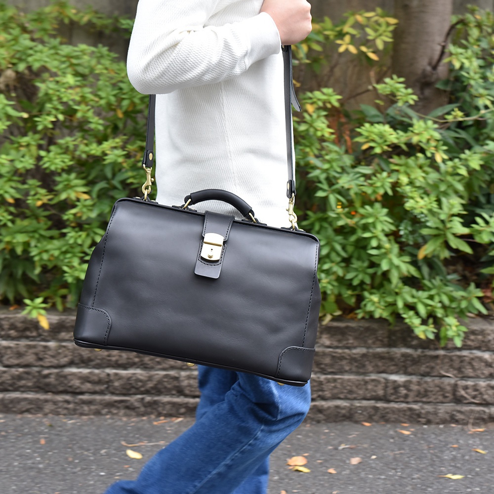 黑色皮革製成的雙用公事包，配有金色鎖扣和提把，可肩背或手提，展現簡約商務風格。