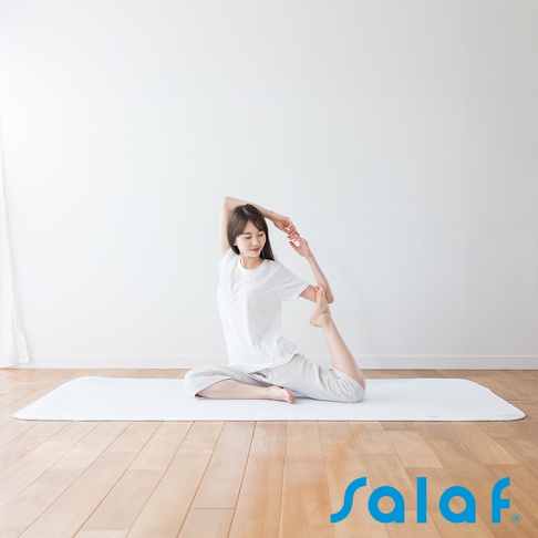 一位身穿白色上衣和灰色長褲的亞洲女性，正在一個白色的 SALAF 涼感睡墊上練習瑜珈。她以單腿盤坐的姿勢，將一隻腳彎曲至身後，並用同側的手抓住腳踝，身體向後仰，展現優美的體態。
