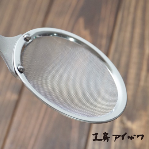 銀色不鏽鋼萬用撈泡勺，帶有細網篩網，勺柄連接處有鉚釘固定，放置於木紋背景上。