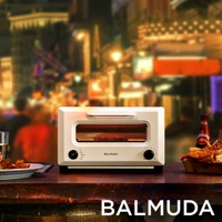 ReBaker 烤麵包機，奶油白色，金屬材質，設有玻璃烤箱門，可見內部正在烘烤的食物。烤麵包機正面有黑色旋鈕和按鈕，以及 BALMUDA 品牌標誌。