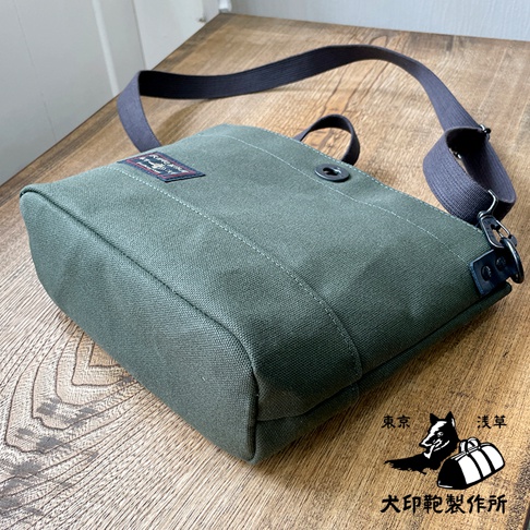 一個綠色的純綿帆布迷你側背包，帶有深灰色織帶和黑色金屬扣件，包包正面有一個品牌標籤。