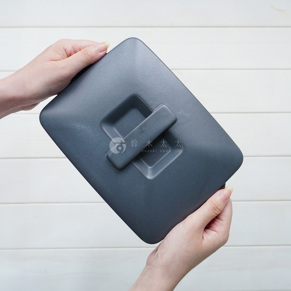 兩隻手拿著一個深灰色、帶有長方形把手的陶製烤盤蓋，烤盤蓋表面光滑，邊緣圓潤。