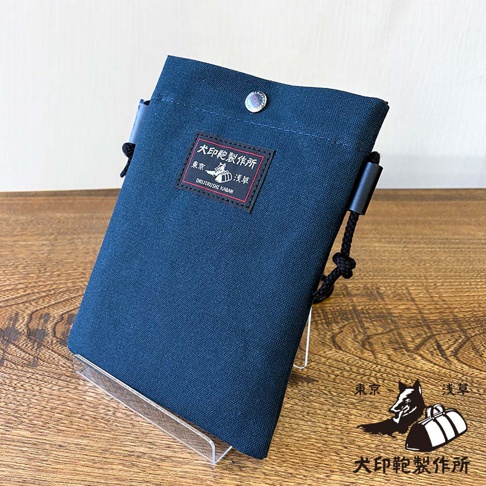 犬印鞄製作所出品的深藍色帆布手機包，上面有日文和英文的品牌標識，包的右側掛著繩子，包包斜放在透明展示架上，背景是木紋桌面。