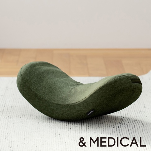 綠色布料製成的U型搖搖枕，表面有織紋，一側有「& MEDICAL」標誌，放在白色地毯上，背景為木地板和白色牆面。