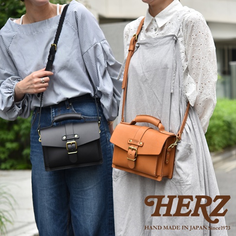 兩款職人手作皮革迷你單肩包，一款為黑色，另一款為棕褐色。兩款包皆為方形設計，配有頂部提把、可調節肩帶、前蓋開合的皮帶與金屬扣環。