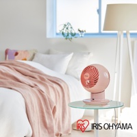 粉紅色圓形空氣循環扇，置於玻璃桌上，背景為臥室，有床鋪、枕頭、毯子和落地燈。