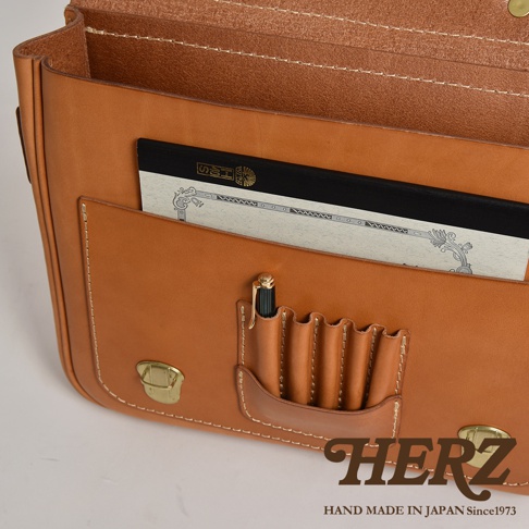 職人手作皮革傳統歐風三用學生包 BC-75，展示包款的駝色皮革，內部有一個筆槽，插著一支黑色與金色的筆，前方外袋內放著一本黑色封面的筆記本，包款有黃銅色金屬扣環，整體以白色背景呈現。