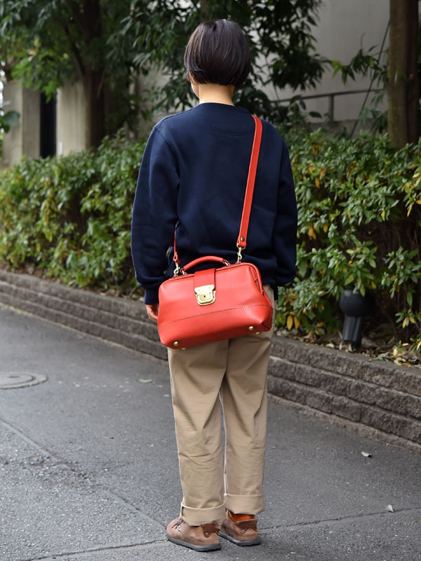 職人手作紅色皮革兩用迷你口金單肩包，帶有金色扣環和鉚釘，斜揹在穿著深藍色衛衣和卡其色長褲的人身上。