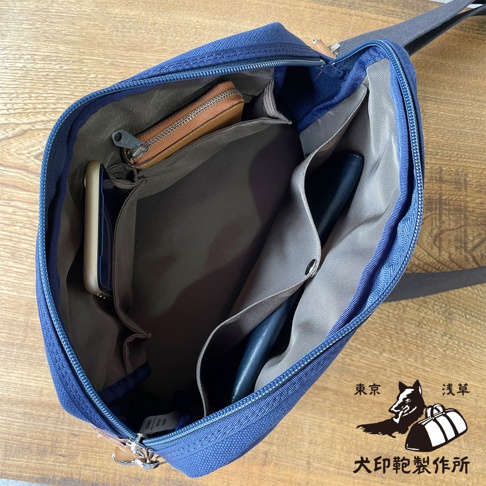 一個打開的藍色石洗帆布旅行側背包，內部有手機、錢包和筆，並印有東京淺草犬印鞄製作所的標誌。