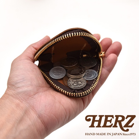 一隻手心托著一個深棕色皮革三角飯糰造型零錢包，拉鍊呈現半開啟狀態，內部裝有數枚日本硬幣。圖片右下方標示著HERZ品牌名稱以及HAND MADE IN JAPAN Since1973字樣。