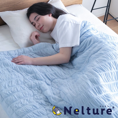 淺藍色 Nelture 單人涼被，表面有細緻的紋理縫線，觸感柔軟，蓋在一位正在床上休息的女性身上。