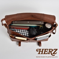 棕色皮革復古郵差包，打開展示內部，包含書籍、紅色皮夾、黑白格紋化妝包、相機、水壺和筆記本。