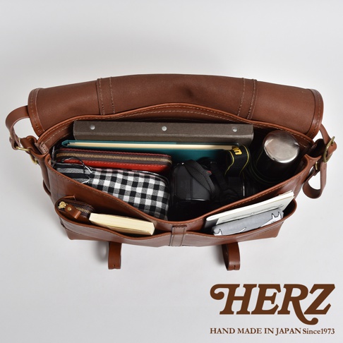 棕色皮革復古郵差包，打開展示內部，包含書籍、紅色皮夾、黑白格紋化妝包、相機、水壺和筆記本。