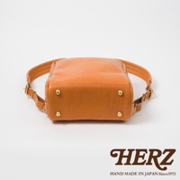 職人手作皮革軟皮商務後背包，採用天然皮革製作，呈現溫潤的橘棕色。包包底部設有四個黃銅色金屬腳釘，增添質感與耐用性。雙肩帶可調節，設計簡潔實用。