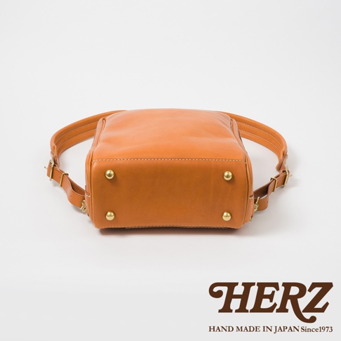 職人手作皮革軟皮商務後背包，採用天然皮革製作，呈現溫潤的橘棕色。包包底部設有四個黃銅色金屬腳釘，增添質感與耐用性。雙肩帶可調節，設計簡潔實用。