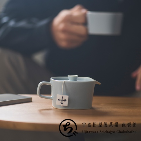 宇治田原製茶場直賣部的 SUKIMA 美濃燒一人用茶壺，淡藍色啞光質感，設計簡約現代，放置於木質桌面上，茶壺側邊掛著帶有十字箭頭標誌的茶包，背景為模糊的人物持杯影像。