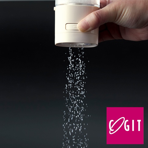 手持一罐米白色塑膠材質的定量灑鹽罐，罐身有按壓按鈕，正定量地灑出細小的白色顆粒，背景為深色。