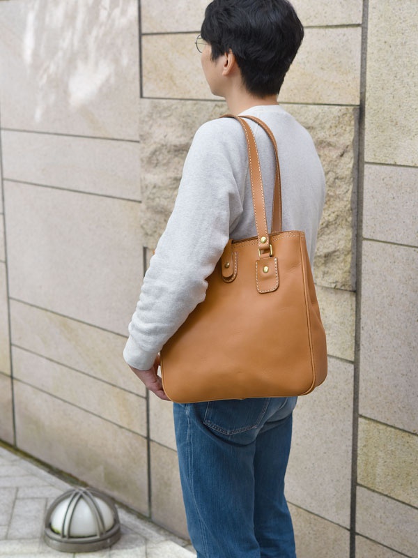 淺棕色皮革托特包，附有原子釦和對比色縫線，由男性背於肩上。