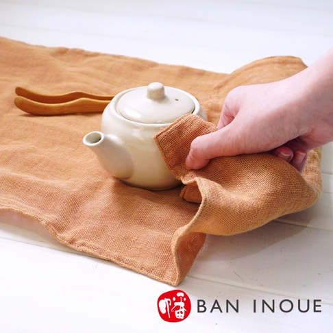 一塊橘棕色、三層蚊帳棉織的廚房拭巾，用手拿取擦拭白色茶壺。