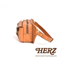 職人手作皮革騎士隨身斜背包，採用天然皮革製成，呈現溫潤的淺棕色。包款側邊帶有拉鍊開口，並以金屬釦件和皮革帶固定，設計簡潔實用。