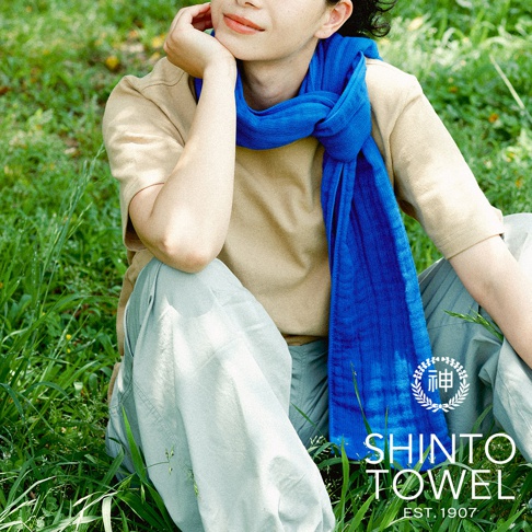 一位人士身穿淺褐色上衣及淺綠色褲子，頸上圍著一條鮮豔的藍色圍巾。圍巾為2.5層紗材質，呈現細緻的織紋，並在胸前打結。背景為綠色草地。