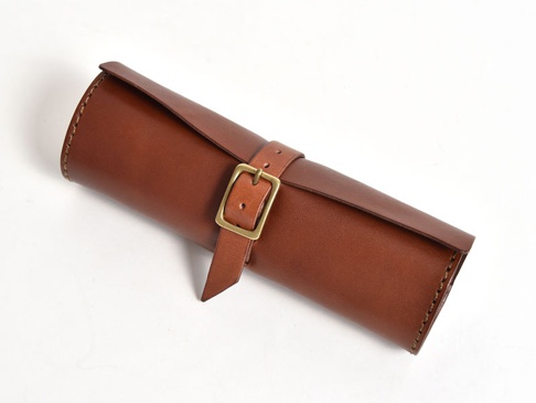 職人手作的棕色皮革捲捲收納筆袋，以皮革捲成圓筒狀，一條皮革繫帶搭配黃銅色扣環固定，邊緣有縫線裝飾。