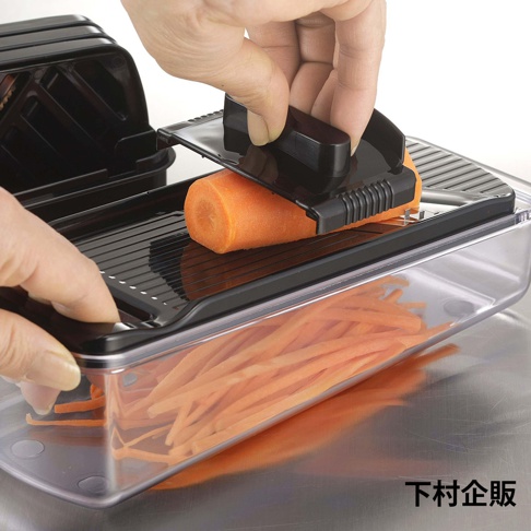 一隻手正使用黑色四合一刨絲器處理紅蘿蔔，刨出的細絲落入下方透明的收納盒中，刨絲器右下角印有下村企販品牌名稱。