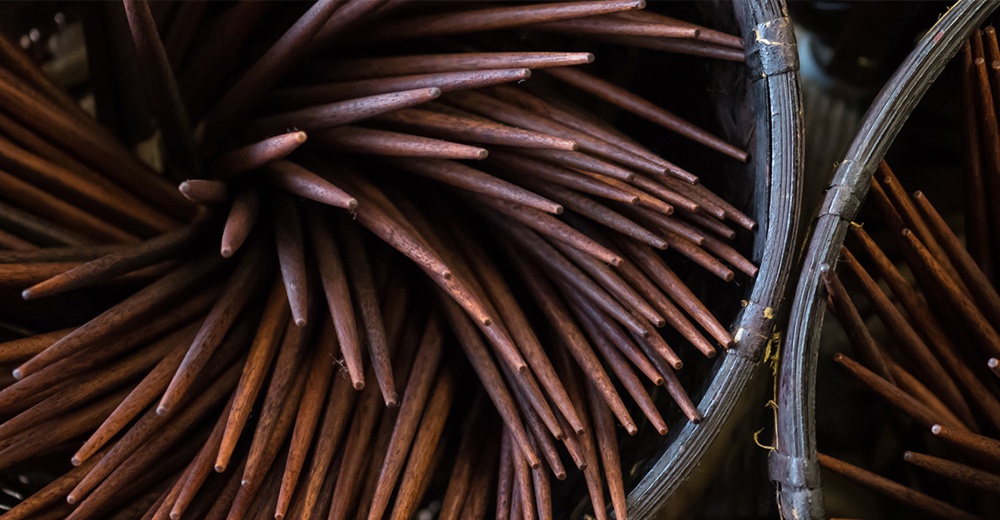 一堆天然原色木筷，呈現深棕色調，筷子末端削尖，材質為木頭，部分露出編織的容器邊緣。