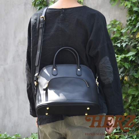 黑色皮革貝殼手提包，設有金色拉鍊和金屬腳釘，可肩背或手提。